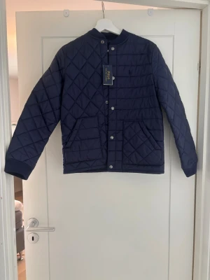 Mörkblå pufferjacka från Polo Ralph Lauren - Snygg mörkblå pufferjacka från Polo Ralph Lauren med quiltat mönster, raka ärmar och två stora fickor framtill. Jackan har tryckknappar och ribbade muddar vid ärmsluten. Perfekt för kalla dagar och ger en stilren look. Storleken är 14-16 år och passar ungefär en S. har aldrig använts, fortfarande med prislapp.