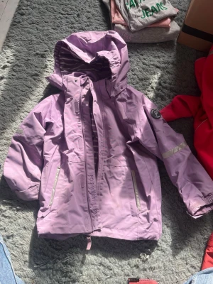 Lila vindjacka för barn från Polarn O. Pyret - Lila vindjacka från Polarn O. Pyret med huva, dragkedja och reflexdetaljer. Jackan har två fickor framtill och justerbar nederkant. Perfekt för blåsiga dagar.