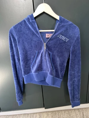 Blå velour hoodie från Juicy Couture - Mysig blå hoodie i velour från Juicy Couture med dragkedja, huva och känguruficka. Broderad JUICY-text framtill och glittrig Juicy Couture-design med stenar på ryggen. Perfekt för en trendig och avslappnad look.