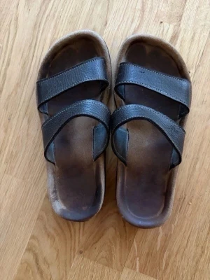 Svarta sandaler med två remmar - Snygga svarta sandaler med två breda remmar i läderimitation och bekväm fotbädd. Sulan är i brunt och svart med en klassisk slip-in design. Perfekta för sommaren och lätta att matcha till olika outfits.