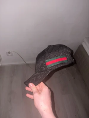 Svart keps - Svart stilren designerkeps med klassiskt GG-monogram över hela kepsen och detaljer i grönt och rött på sidorna. Kepsen har böjd skärm, justerbar passform och en exklusiv look som passar både till vardags och streetwear-outfits. Elegant mörk design som är lätt att matcha med olika kläder och ger en lyxig känsla.