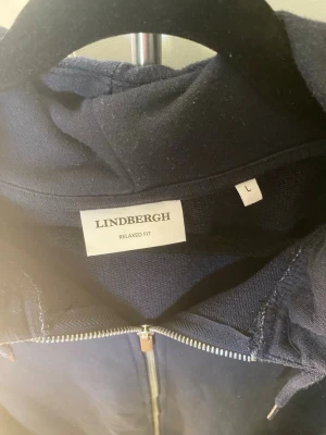 Mörkblå hoodie från Lindbergh - Mörkblå hoodie från Lindbergh i relaxed fit med dragkedja framtill och justerbar huva. Tillverkad i mjukt material, perfekt för en chill och avslappnad stil. Klassisk design med ribbade muddar och fickor på sidorna.