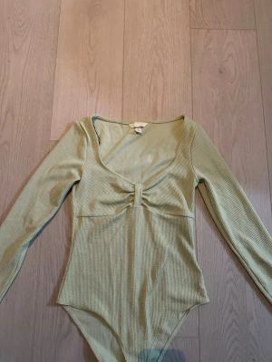 Ljusgrön ribbad långärmad body - Trendig ljusgrön body med lång ärm och ribbad struktur. Djup rundad urringning och snygg rynkdetalj framtill. Perfekt att styla till jeans eller kjol för en cool look.