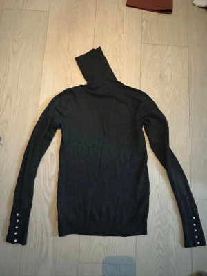 Svart polotröja med pärlknappar - Säljer en svart stickad polotröja med lång ärm och snygga pärlknappar vid ärmsluten. Tröjan har en tight passform och är perfekt för lager på lager. Enkel men elegant design som passar till mycket.