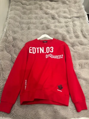 Röd sweatshirt från Dsquared2 - Röd sweatshirt från Dsquared2 med tryckta vita och svarta texter både fram och bak. Rund halsringning, långa ärmar och ribbade muddar. Snygg detalj med lönnlöv och märkets logga framtill. Perfekt för en sportig och trendig look.