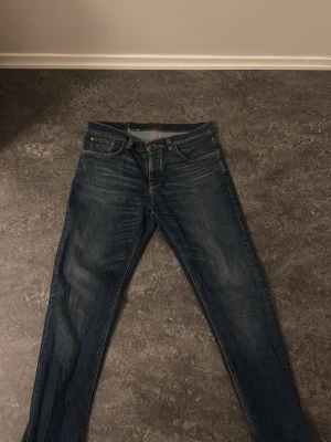Mörkblå jeans från Nudie Jeans - Säljer ett par mörkblå jeans från Nudie Jeans med klassisk femficksmodell och orangea sömmar. Jeansen har en smal passform och är tillverkade i denim med lätt tvättad look. Perfekta för en stilren vardagsoutfit.