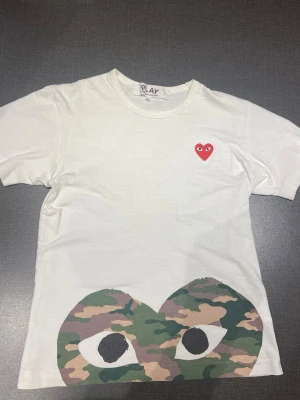 Vit Comme des Garçons Play t-shirt - Vit t-shirt från Comme des Garçons Play med det ikoniska röda hjärtat med ögon på bröstet och ett stort camo-mönstrat hjärta med ögon nedtill. Rund halsringning och korta ärmar. Perfekt nu inför sommaren. Pris kan diskuteras. Hör av er vid frågor.