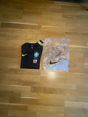 Svart Brasilien Nike träningströja - Snygg svart träningströja från Nike med Brasilien-emblem på bröstet och gula samt orangea loggor på ärmarna. Tröjan har halv dragkedja framtill och är tillverkad i ett funktionsmaterial som passar perfekt till träning. Officiell landslagsmodell.