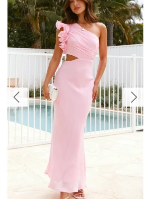 Ljusrosa one shoulder långklänning - Ny ljusrosa långklänning med prislapp kvar. Den har en one shoulder-design och volangdetalj på axeln. Klänningen har en cut-out vid midjan och är figurnära med en mjuk, flytande siluett. Perfekt för speciella tillfällen och ger en romantisk look.