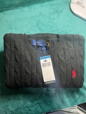 Mörkgrå kabelstickad tröja Polo Ralph Lauren - Kabelstickad mörkgrå tröja från Polo Ralph Lauren med klassisk röd logga broderad på bröstet. Tröjan har rund halsringning och långa ärmar. Perfekt för dig som gillar stilrena och tidlösa plagg.