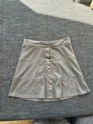 Grå kortkjol med knappar framtill - Snygg grå kortkjol i mjukt material med dekorativa knappar framtill. Kjolen har en lätt utsvängd form och bred linning med resår  för extra komfort. Perfekt att matcha med en enkel topp eller stickad tröja.