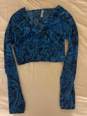 Blå croppad långärmad topp med zebramönster - Snygg croppad topp i blått och svart zebramönster. Långärmad modell med v-ringning och hakknäppning framtill. Tunn och stretchig polyester som sitter tight och ger en trendig look.