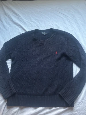 Mörkblå kabelstickad tröja Polo Ralph Lauren - Mörkblå kabelstickad tröja från Polo Ralph Lauren med rund halsringning och röd broderad logga på bröstet. Tröjan har långa ärmar och breda ribbade muddar vid ärmslut och nederkant. Perfekt för en stilren och klassisk look. Passar en 12-13 åring 
