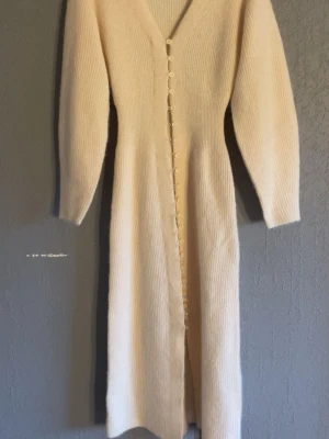 Lång stickad klänning med knappar - Lång, ribbstickad klänning i ljus beige med v-ringning och långa ärmar. Klänningen har knappar framtill hela vägen ner och är tillverkad i ett mjukt stickat material. Perfekt för en mysig och stilren look.