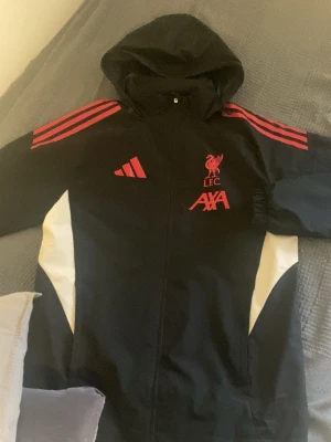 Liverpool FC vindjacka från Adidas - Snygg svart vindjacka från Adidas med röda detaljer och Liverpool FC-logga på bröstet. Jackan har huva, dragkedja framtill och vita paneler på sidorna. Använd 5-6 gånger 