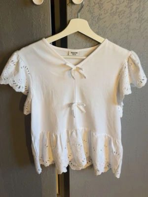 Vit blus med broderade volanger Kappahl - Söt vit blus från Kappahl med broderade volangdetaljer på ärmar och nederkant. Toppen har v-ringning med två knytband framtill och korta ärmar. Perfekt för en fräsch och somrig look.