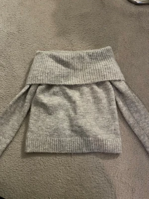 Grå off shoulder stickad tröja - Säljer en snygg grå stickad tröja med bred off shoulder-krage och långa ärmar. Tröjan har ribbstickade detaljer vid krage, ärmslut och nederkant. Perfekt för dig som vill ha en trendig och mysig look.
