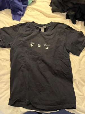 Svart Off-White t-shirt med tryck - Svart t-shirt från Off-White med vit logga och symboler på bröstet samt stort blått och vitt grafiskt tryck på ryggen. Klassisk passform och rund halsringning. Perfekt för dig som gillar streetwear och unika prints. Inte @kta men snygg ändå! 🙃 står storlek M men sitter som en xs eller S 