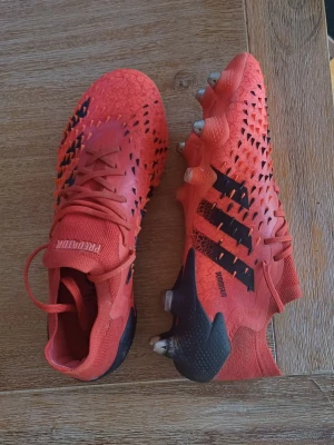 Röda Adidas Predator fotbollsskor - Säljer ett par röda Adidas Predator fotbollsskor med svarta detaljer och klassiska tre ränder på sidan. Skorna har snörning, taggig textur på ovandelen för bättre bollkontroll och dobbar för spel på gräs. Skorna är till AG, storlek 40,5. Skorna har bara använts ett fåtal gånger dom är I mycket bra skick. Skorna är elite.