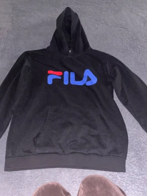 Svart hoodie från FILA med logga - Svart hoodie från FILA med stor blå och röd logga på bröstet. Klassisk känguruficka framtill och ribbade muddar vid ärmslut och nederkant. Tillverkad i mjukt bomullsmaterial och har en skön huva. Perfekt för dig som gillar sportig och avslappnad stil.