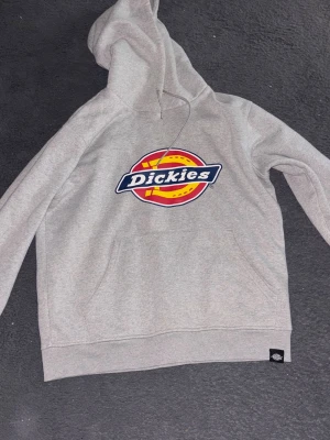 Grå Dickies hoodie med logga - Snygg grå hoodie från Dickies med stor färgglad logga på bröstet. Klassisk känguruficka framtill och justerbar huva med dragsko. Mjuk bomullsblandning som är skön att ha på sig. Perfekt för en avslappnad streetwear-look. ANTVÄNT 2-3 gånger 