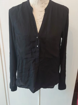 Svart blus med v-ringning - Stilren svart blus med v-ringning och bröstficka. Blusen har långa ärmar och knäppning framtill med små knappar. Tunt och lätt material som ger en avslappnad känsla. Ärm kan vikas upp med knapp till trekvartsärm.