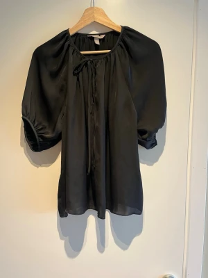 Svart blus med puffärm och knyt - Snygg svart blus i glansigt material med korta puffärmar och knytband vid halsen. Blusen har en avslappnad passform och rundad halsringning. 