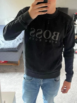 Svart sweatshirt från Hugo Boss - Svart sweatshirt från Hugo Boss med stor BOSS-logga framtill. Tröjan har rund halsringning, ribbade muddar och är tillverkad i mjukt material. Perfekt för en stilren och avslappnad look.