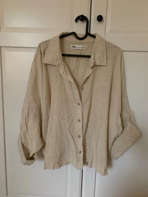 Beige randig skjorta från Zara - Snygg beige skjorta i linne från Zara med tunna vita ränder. Skjortan har klassisk krage, knäppning framtill och långa ärmar som kan vikas upp med knapp.