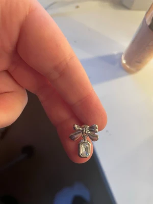 Söta silverfärgade rosettörhängen - Ett par örhängen i silverfärg med rosettformad detalj och en rektangulär klar sten som hänger nedanför. Perfekt accessoar för att ge en gullig och elegant touch till din stil.