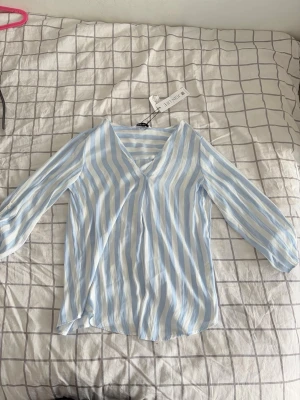 Somrig blus - Snygg blus med blå och vita vertikala ränder. Blusen har v-ringning. Tillverkad i ett lätt och luftigt material som känns skönt mot huden. Perfekt för en fräsch och stilren look.