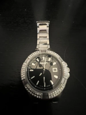 Silverfärgad klocka från GANT - Stilren klocka från Gant med silverfärgat armband i metall och svart urtavla. Runt boetten sitter glittrande stenar som ger extra bling. Liten storlek. Länkar medföljer ej. Batteri behövs!