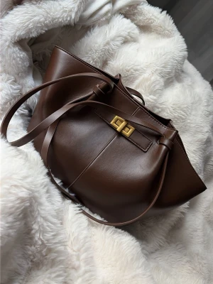 Brun handväska i läder med guldspänne - Snygg brun handväska i slätt läder med långa axelband och elegant guldspänne framtill. Väskan har en mjuk, rundad form och minimalistisk design som ger en stilren look. Den är helt felfri och har inga skador🫶 Köptes på Gina tricot för 399kr, Den har även 3 innerfickor varav en har dragkedja och de andra är öppna