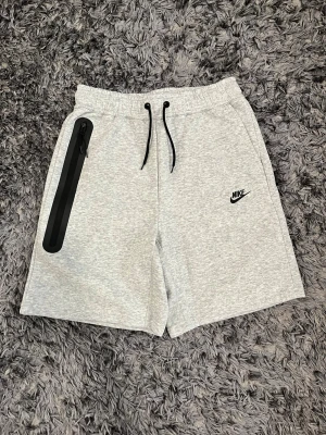 Helt nya grå Nike tech shorts med dragkedja - Snygga ljusgrå shorts från Nike med svart logga på vänster ben. Shortsen har elastisk midja med svart snörning och en praktisk ficka med svart dragkedja på höger sida. Perfekta för träning eller chill. Mjukt material som känns skönt mot huden.