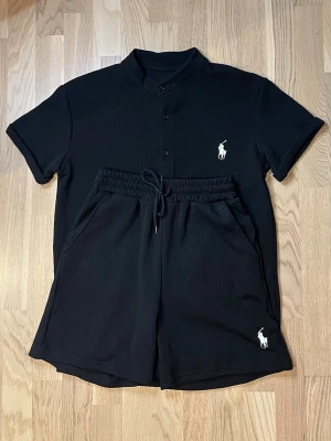 Svart set från Ralph Lauren - Snyggt svart set från Ralph Lauren med kortärmad skjorta och matchande shorts. Skjortan har knappar framtill och klassisk krage, båda plaggen har den ikoniska vita polospelaren broderad. Materialet ser mjukt och bekvämt ut, perfekt för en avslappnad look. Skriv vid fler frågor🙌