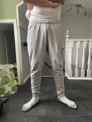 Grå mjukisbyxor från Nike - Snygga grå mjukisbyxor från Nike med smal passform och resår i midjan. Byxorna har en diskret Nike-logga på vänster ben och är tillverkade i mjukt material som är skönt att ha på sig. Perfekta för en avslappnad stil.