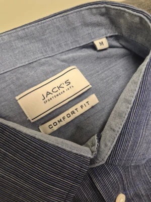 Blå randig skjorta från Jack's comfort fit M - Snygg blå skjorta från Jack's med tunna vita ränder och klassisk krage. Skjortan har lång ärm, vita knappar och en relaxed comfort fit. Perfekt för dig som gillar en stilren och enkel look.