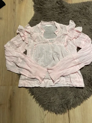 Ljusrosa blus med volanger - Söt ljusrosa blus med volangdetaljer på axlarna och spetsinslag. Blusen har långärmade, lätt puffade ärmar och är gjord i ett tunt, luftigt tyg. Perfekt för dig som gillar en feminin och trendig stil. Aldrig använd