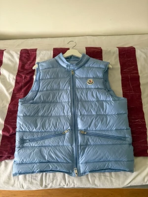 Ljusblå dunväst från Moncler - Snygg ljusblå dunväst från Moncler med klassisk logotyp på bröstet. Västen har två snedställda dragkedjefickor. Perfekt för lager-på-lager och ger en clean look. Tillverkad i glansigt material med quiltad design. NFT tag se sista bild 