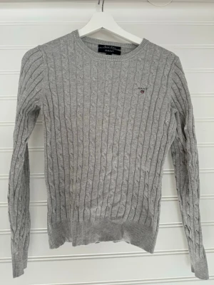 Grå kabelstickad tröja från GANT - Kabelstickad grå tröja från GANT med rund halsringning och diskret logga broderad på bröstet. Tröjan har långa ärmar och ribbade muddar vid ärmslut och nederkant. Perfekt för lager-på-lager och en klassisk stil.