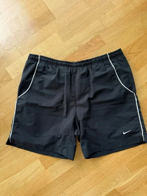 Svarta träningsshorts från Nike - Svarta träningsshorts från Nike med vit piping längs sidorna och liten vit logga på vänster ben. Shortsen har elastisk midja och är tillverkade i ett lätt och snabbtorkande material, perfekta för sport och aktivitet.