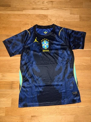 Blå Brasilien tränings t-shirt Nike Brazil  - Snygg blå tränings t-shirt från Nike med Brasilien CBF-logga och gula detaljer. Tröjan har korta ärmar, svart och blått mönster samt meshpaneler i ljusblått på sidorna. Perfekt för fotbollsträning eller gymmet.
