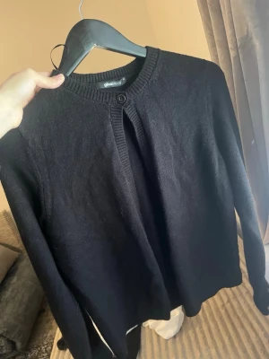 Svart kofta från Gina Tricot - Klassisk svart kofta från Gina Tricot med rund halsringning och knapp framtill. Koftan har långa ärmar och ribbade muddar vid hals, ärmslut och nederkant. Perfekt att slänga över en topp för en enkel och clean look.
