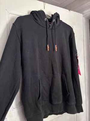 Svart hoodie från Alpha Industries - Snygg svart hoodie från Alpha Industries med klassisk känguruficka och huva med dragsko. Detaljer i form av metallöljetter och bruna snörstoppar. Mjuk bomullskänsla och ribbade muddar. Liten ficka med dragkedja på ena ärmen för extra stil.