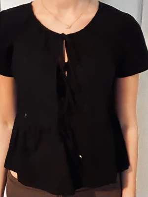 Svart blus med knytning framtill - Svart blus med korta ärmar och knytning framtill. Blusen har en rund halsringning och lätt utställd passform nedtill. Perfekt för en trendig och avslappnad look.