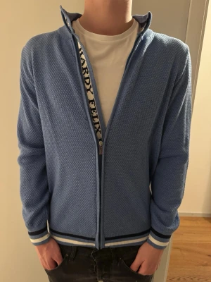 Blå ziptröja från Felix Hardy - Snygg blå ziptröja från Felix Hardy med diskret strukturmönster. Tröjan har vita och mörkblå ränder på muddar och nederkant samt dragkedja framtill. Perfekt för en avslappnad och stilren look.