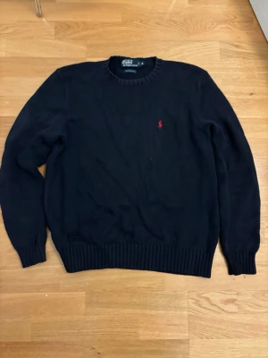 Mörkblå stickad tröja från Polo Ralph Lauren - Klassisk mörkblå stickad tröja från Polo Ralph Lauren med rund halsringning och röd broderad logga på bröstet. Tröjan har ribbade muddar vid ärmslut och nederkant. Perfekt till jeans eller chinos för en clean look.