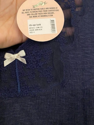 Mörkblå ribbad linnetopp Odd Molly XL - Supersöt mörkblå ribbad linnetopp från Odd Molly med spetsdetaljer och liten rosett framtill. Ärmlös modell med vacker kant nertill och vid halsen. Perfekt att styla med jeans eller kjol för en trendig look.