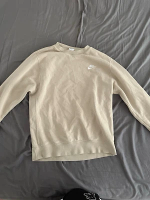 Beige nike sweatshirt  - Snygg beige sweatshirt från Nike med klassisk crewneck och diskret vitt Nike-logga broderad på bröstet. Tröjan har en avslappnad passform och är tillverkad i mjukt material, perfekt för en chill och stilren look.