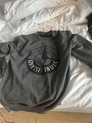 Grå sweatshirt från Stone Island - Snygg grå sweatshirt från Stone Island med stort svart logotryck på bröstet. Tröjan har rund halsringning och långa ärmar. Perfekt för en avslappnad och trendig stil. OBS den är orginelt i storlek L men förminskades i tvätten och passar nu storlek S, (175~180)